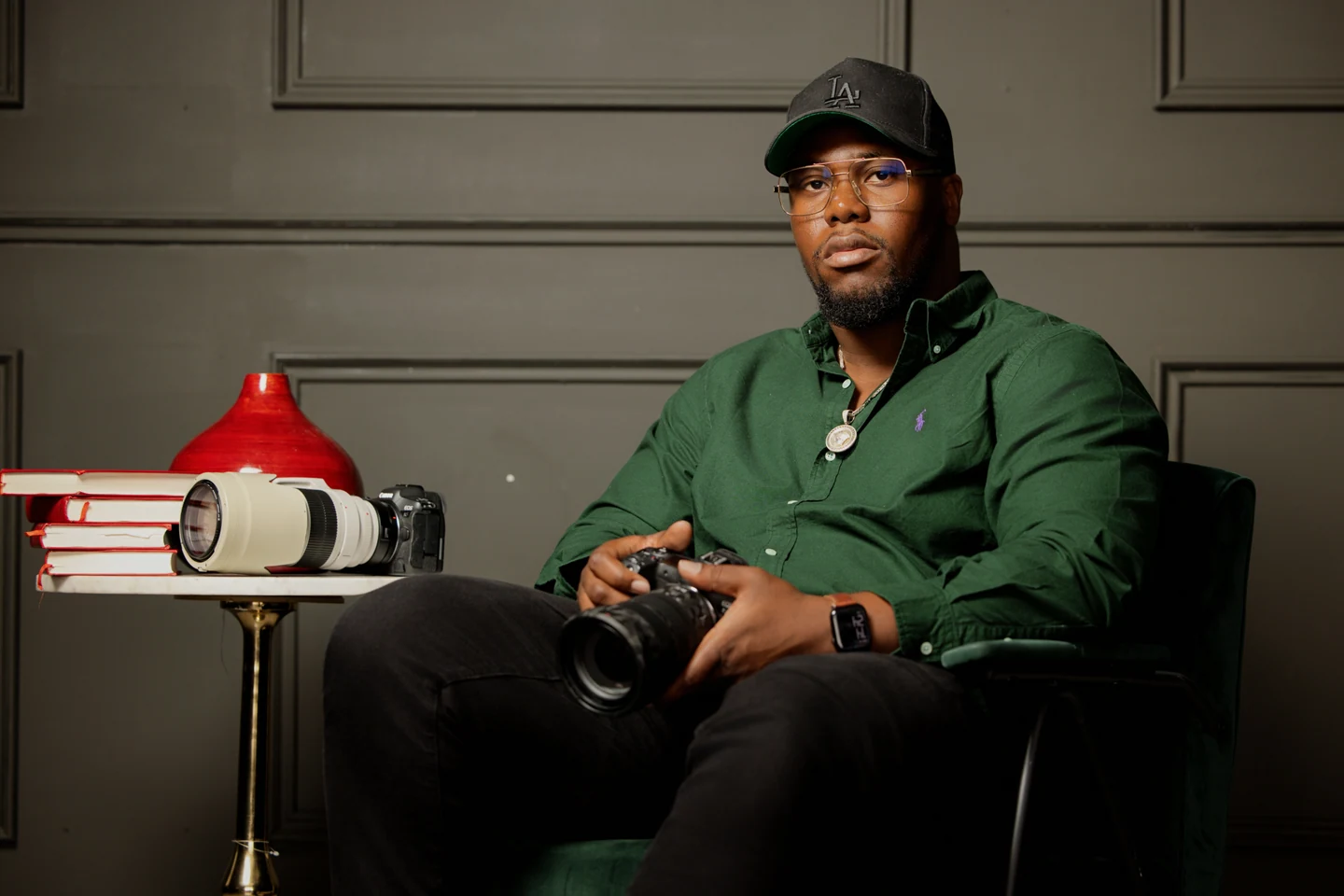 Kalam assis dans un fauteuil, en chemise verte, casquette noire et lunettes, regardant l’objectif avec un appareil photo entre les mains.