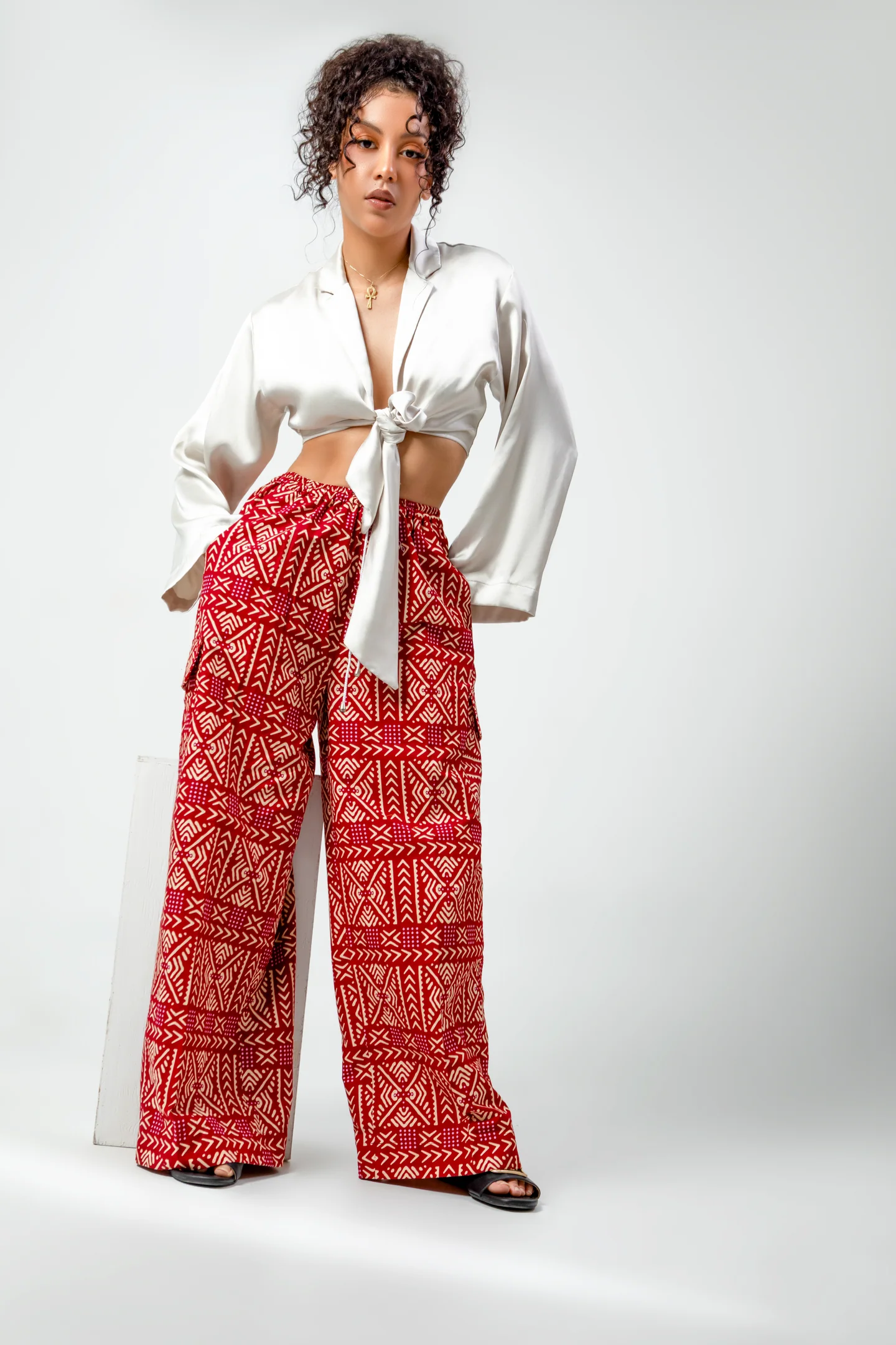 Femme en chemisier blanc noué et pantalon rouge à motifs, debout sur fond clair.