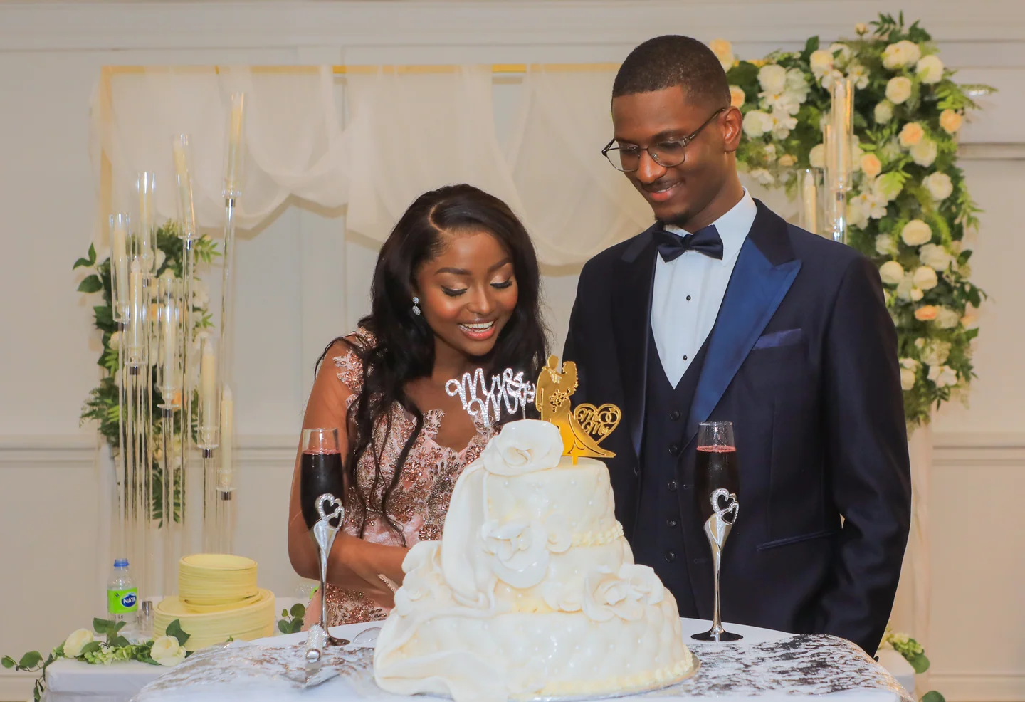 Couple souriant derrière un gâteau de mariage blanc, avec des fleurs et des bougies autour de la table.