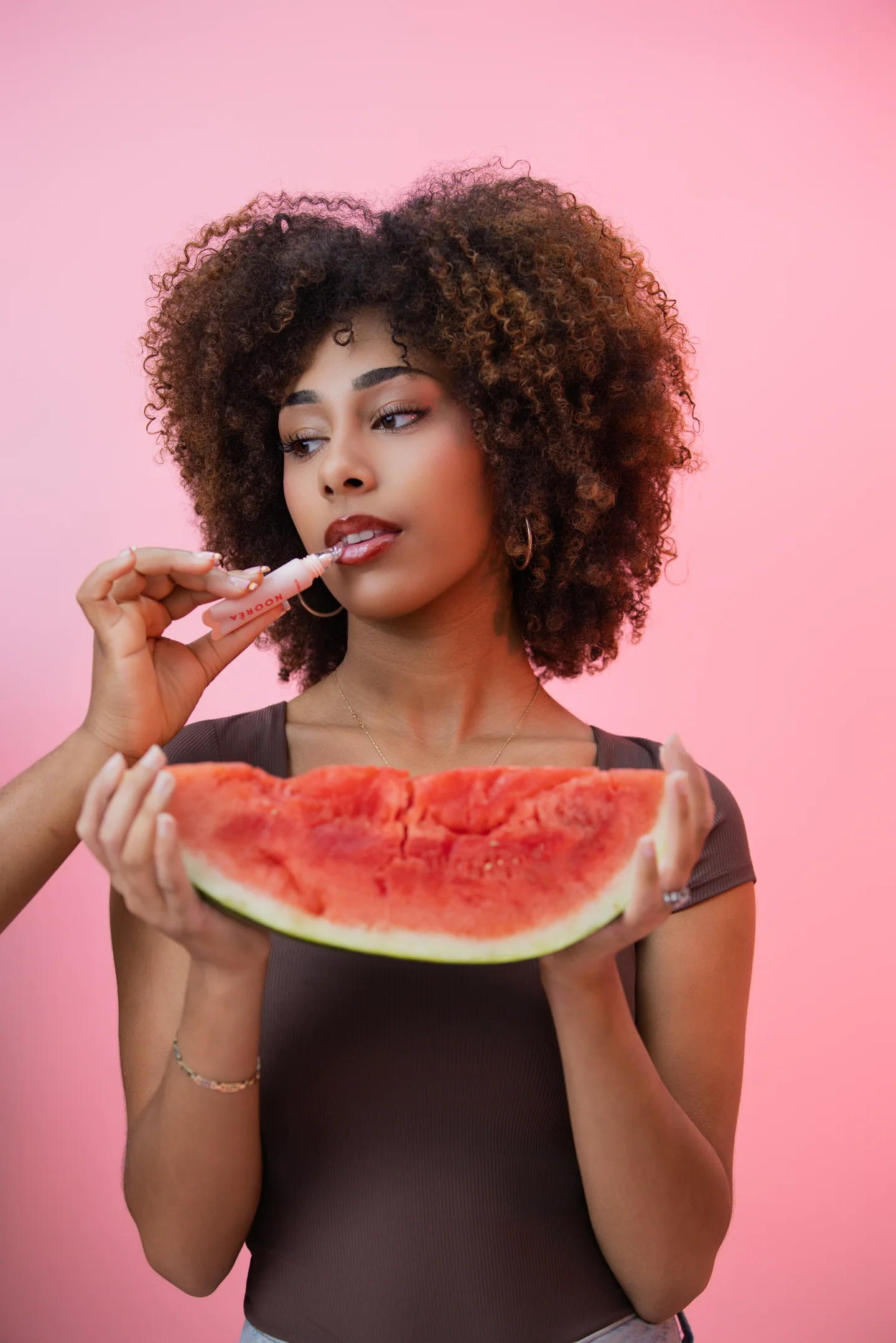 Femme aux cheveux bouclés tenant une tranche de pastèque tout en appliquant du gloss, sur fond rose.