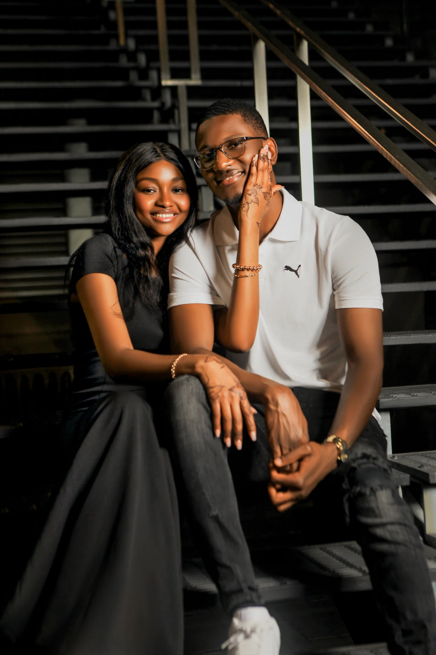 Couple assis dans un escalier sombre, la femme en robe noire posant une main sur la joue de l’homme.