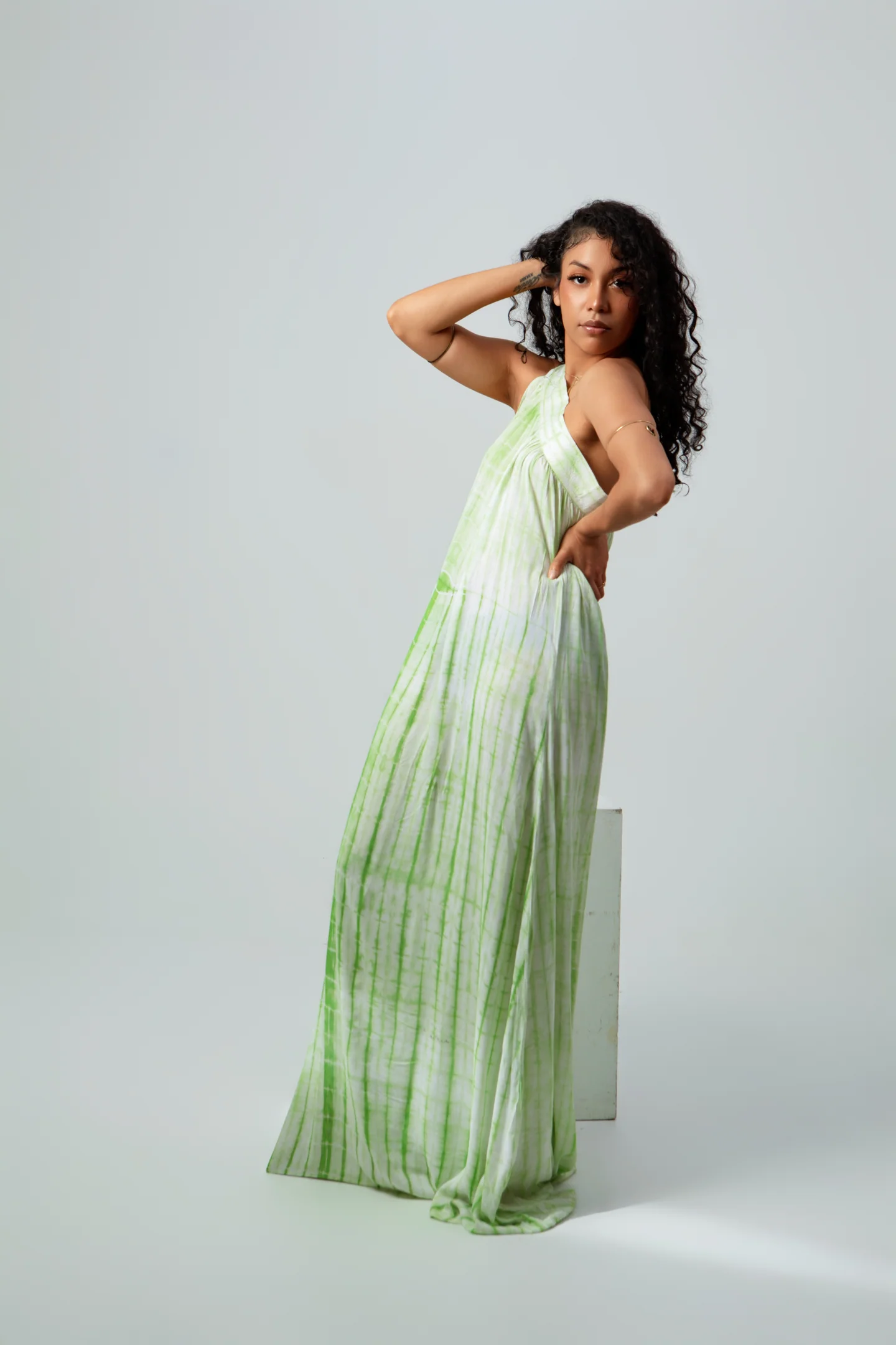 Femme en longue robe vert pâle, une main dans les cheveux et l’autre à la taille, sur fond gris.