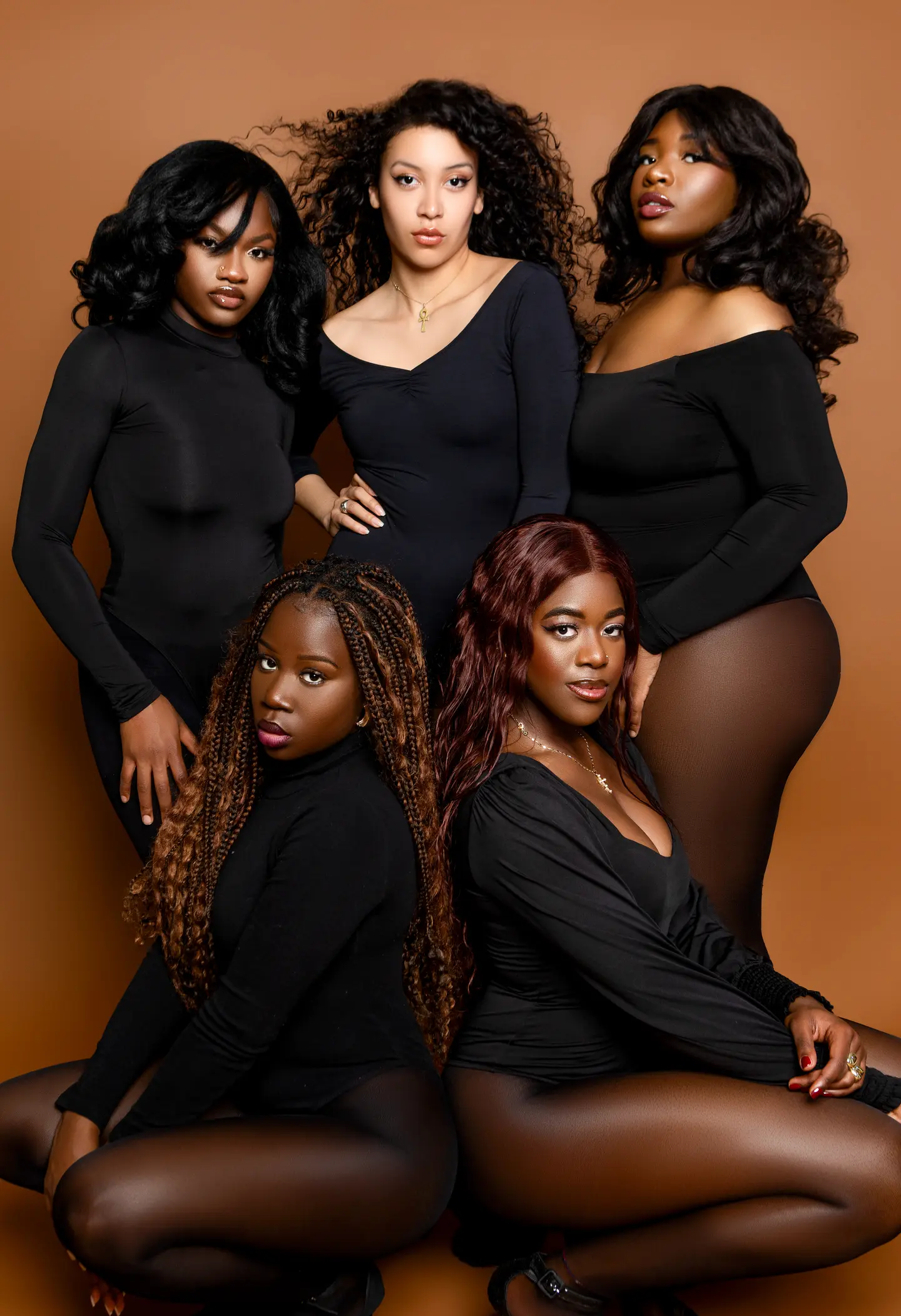 Cinq femmes en tenues noires composent un portrait de groupe sur un fond brun chaleureux.
