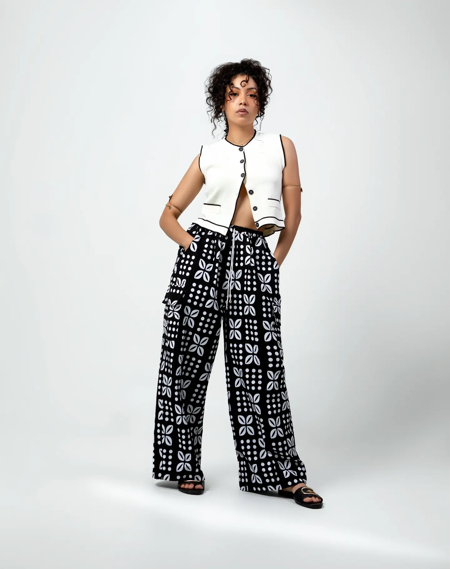 Femme en gilet blanc et pantalon noir à motifs, debout face à l’objectif sur un fond clair.