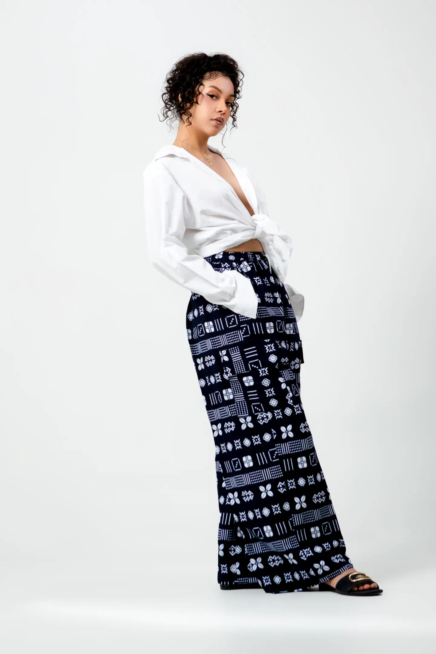 Femme en chemisier blanc et pantalon ample noir à motifs, debout dans une pose souple.