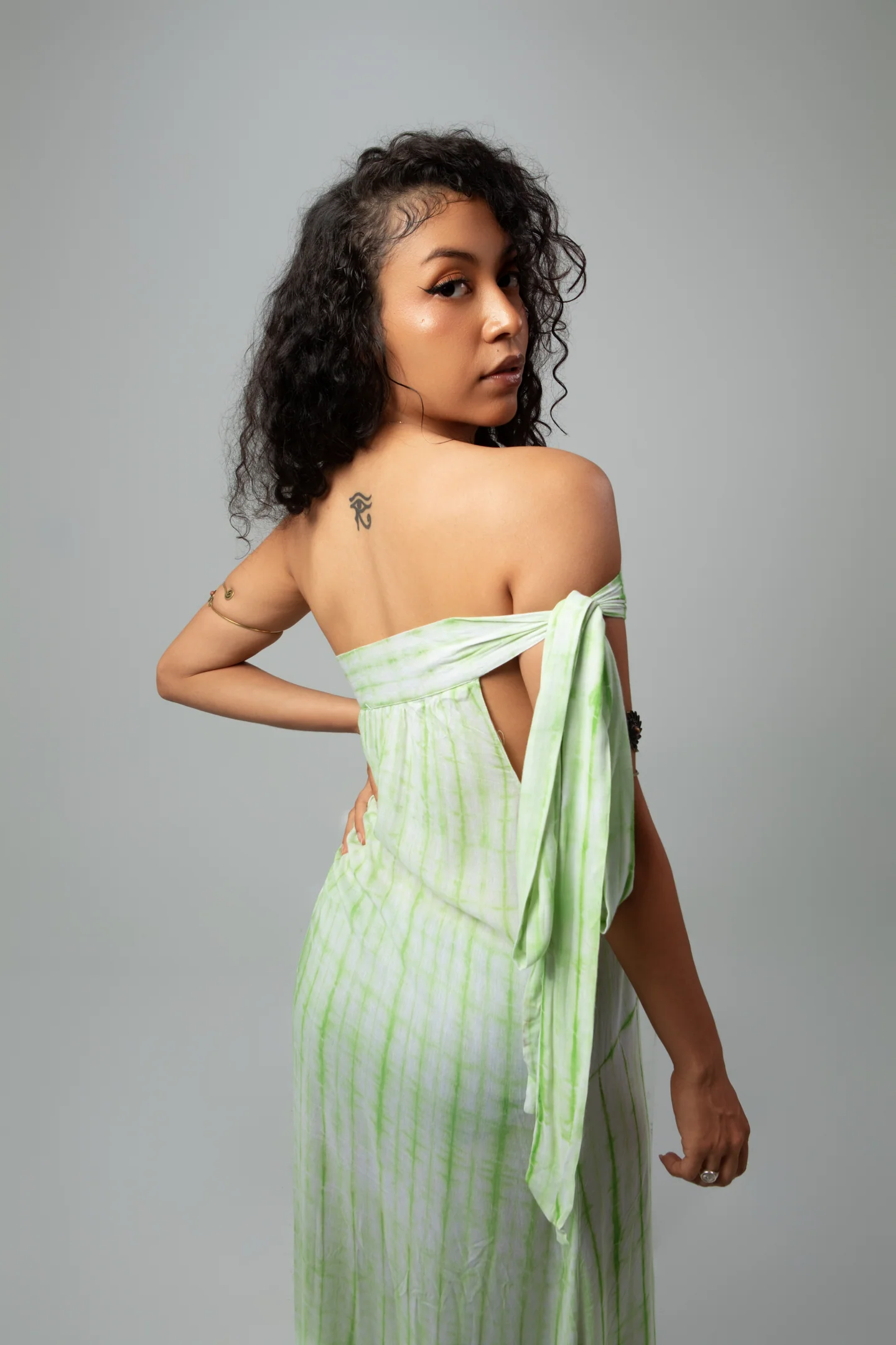 Femme en robe vert pâle vue de dos, tournant le regard vers l’objectif, sur fond gris.
