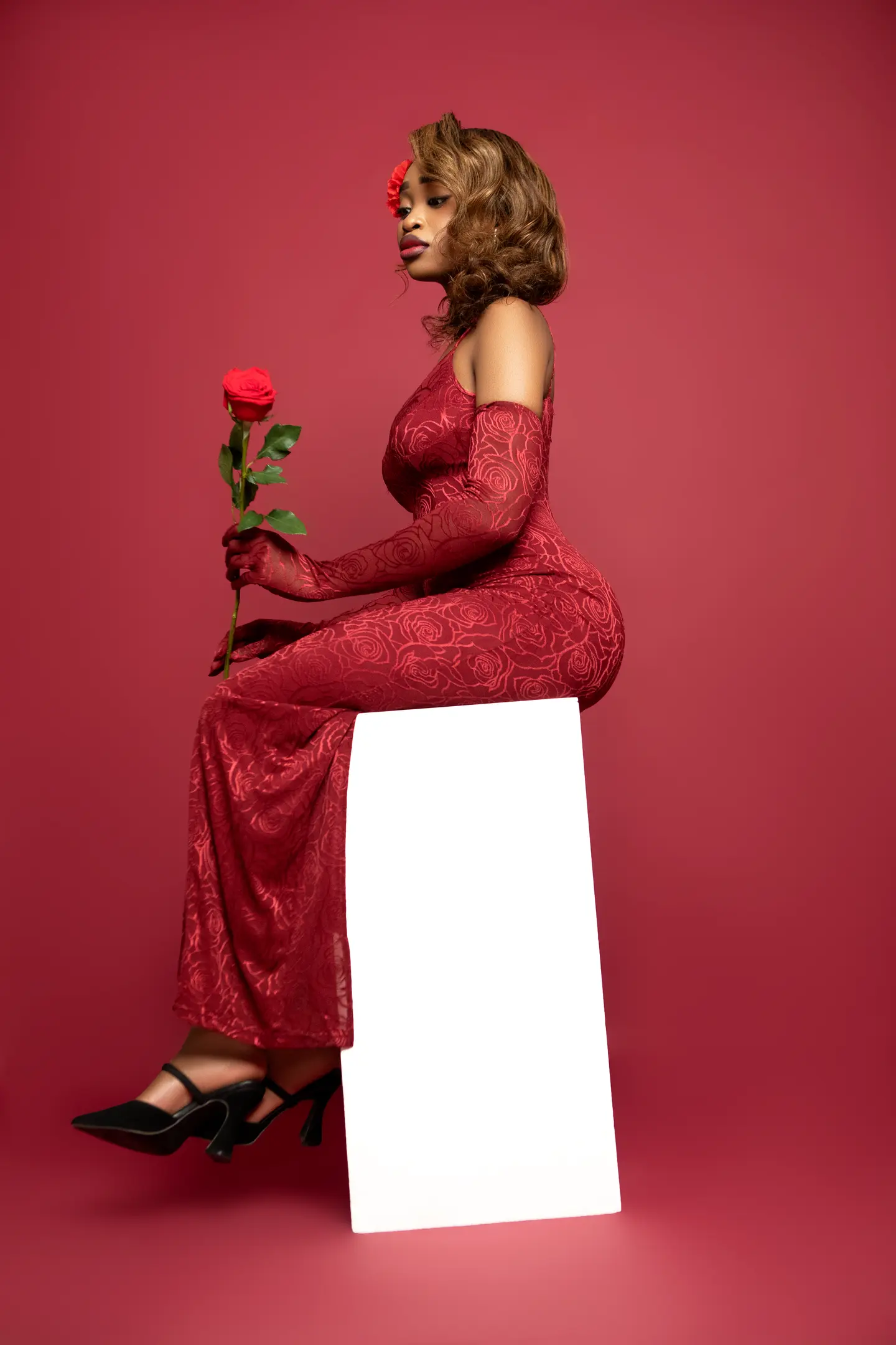 Portrait de mode de profil d’une femme en robe rouge tenant une rose, assise devant un fond carmin.