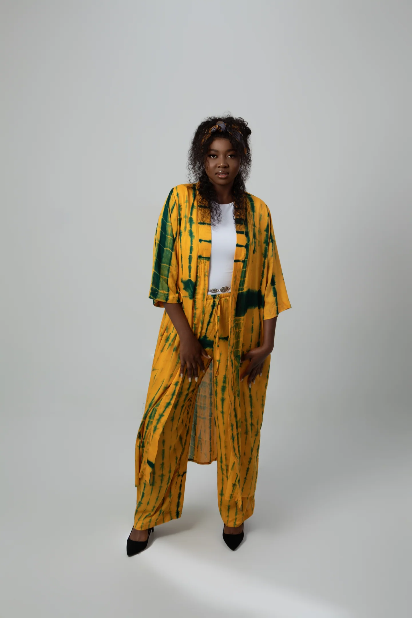 Femme photographiée en pied, portant un ensemble ample jaune et vert sur fond gris clair.