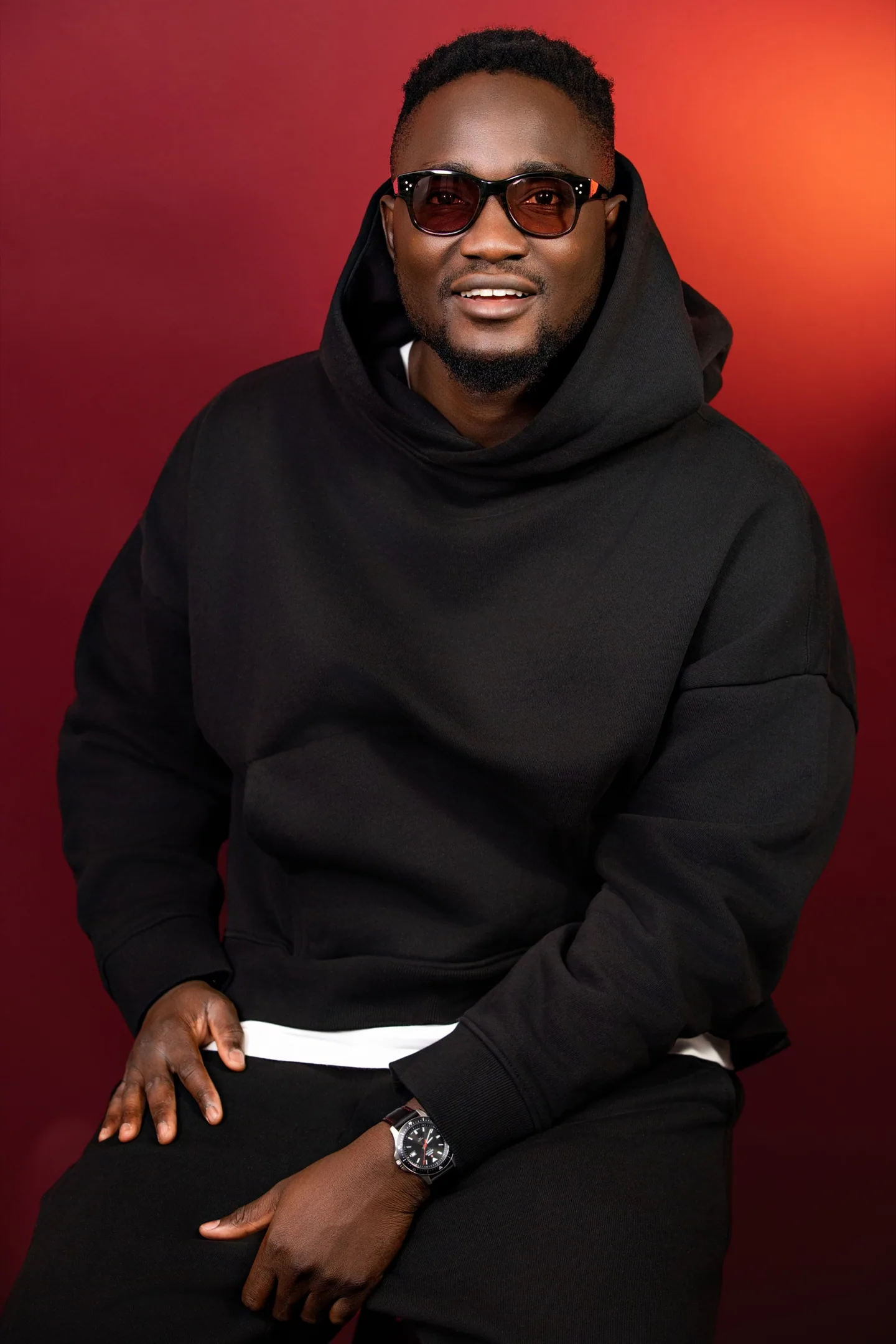 Portrait d’un homme souriant portant des lunettes teintées et un hoodie noir, sur fond rouge.