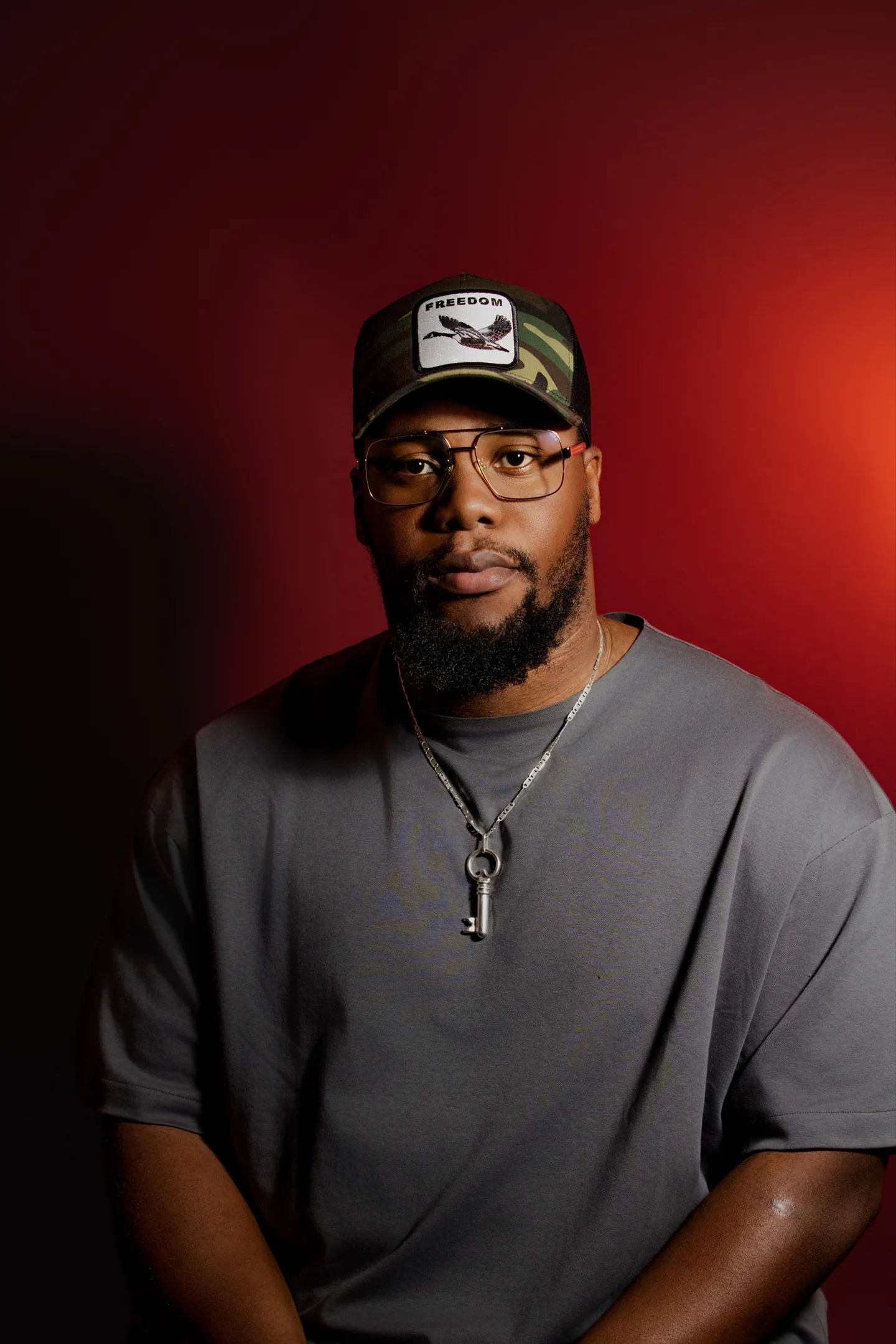 Homme portant une casquette camouflage, des lunettes et un pendentif, photographié sur fond rouge.