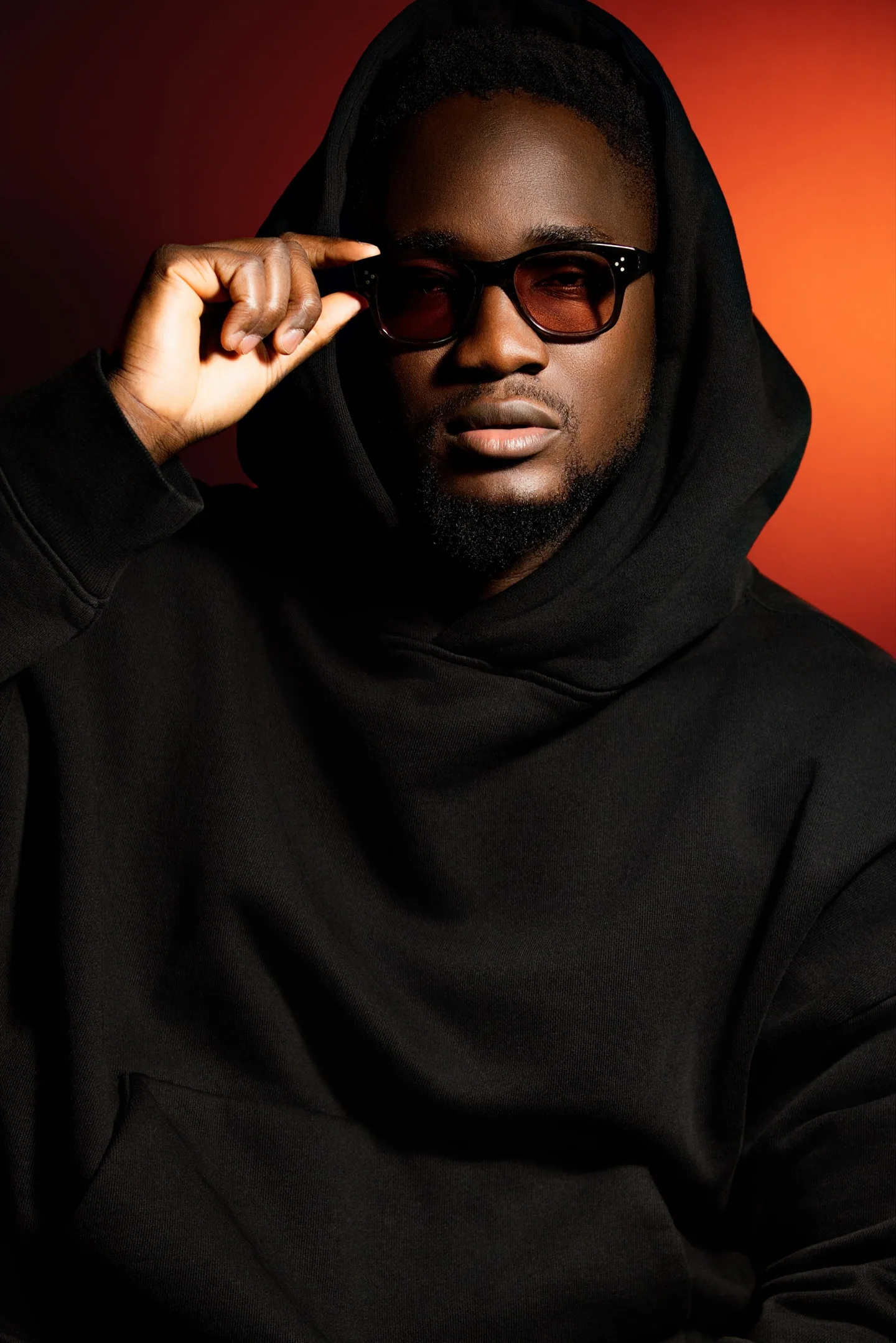 Portrait d’un homme en hoodie noir ajustant ses lunettes, devant un fond rouge.