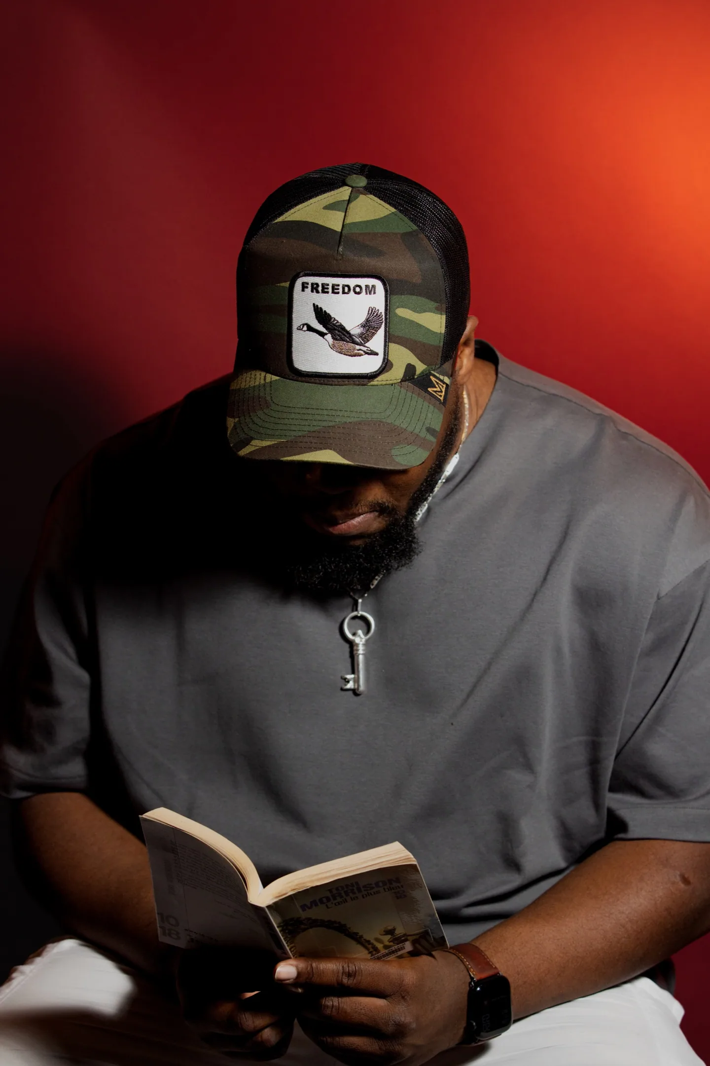 Homme portant une casquette et un haut gris, lisant un livre devant un fond rouge.