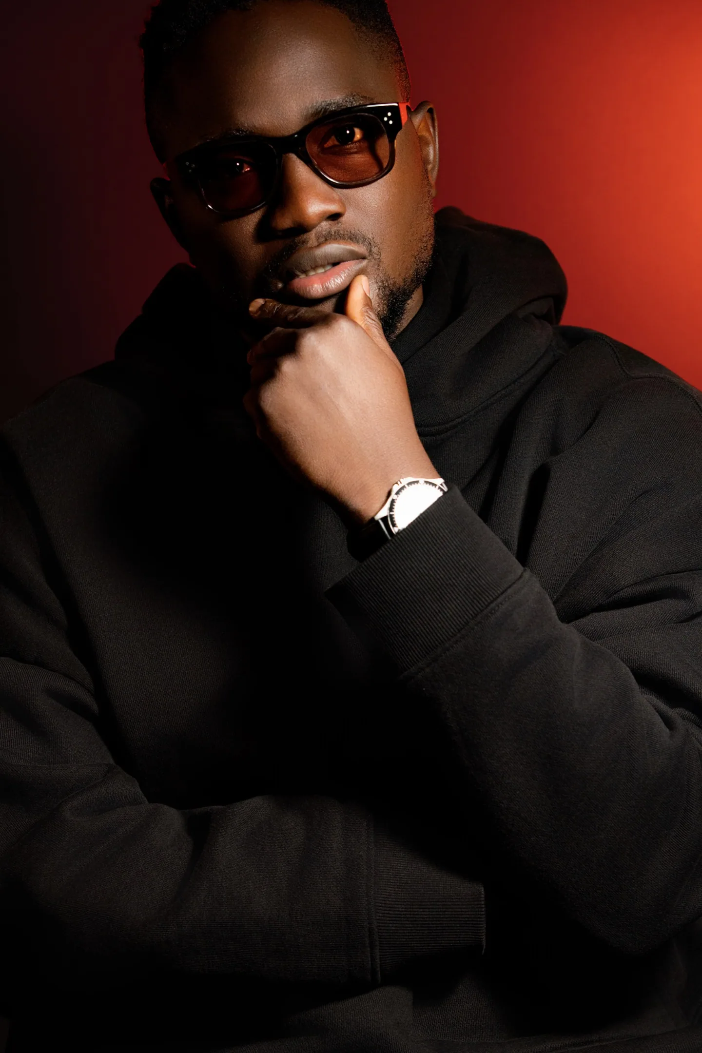 Portrait rapproché d’un homme en hoodie noir et lunettes teintées, éclairé sur fond rouge.