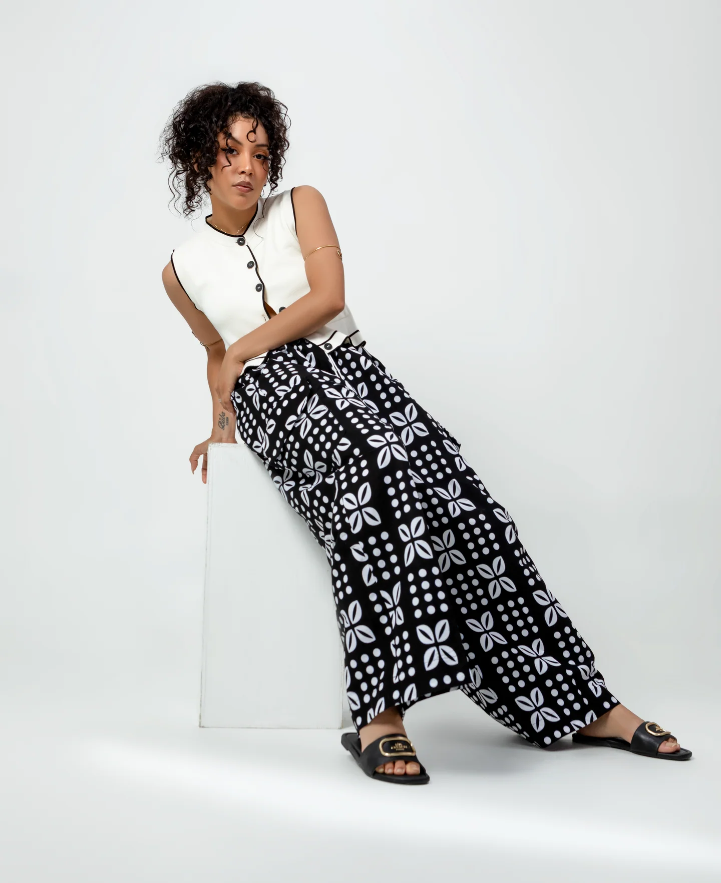 Femme en gilet blanc et pantalon ample noir à motifs blancs, assise de biais sur un cube blanc.