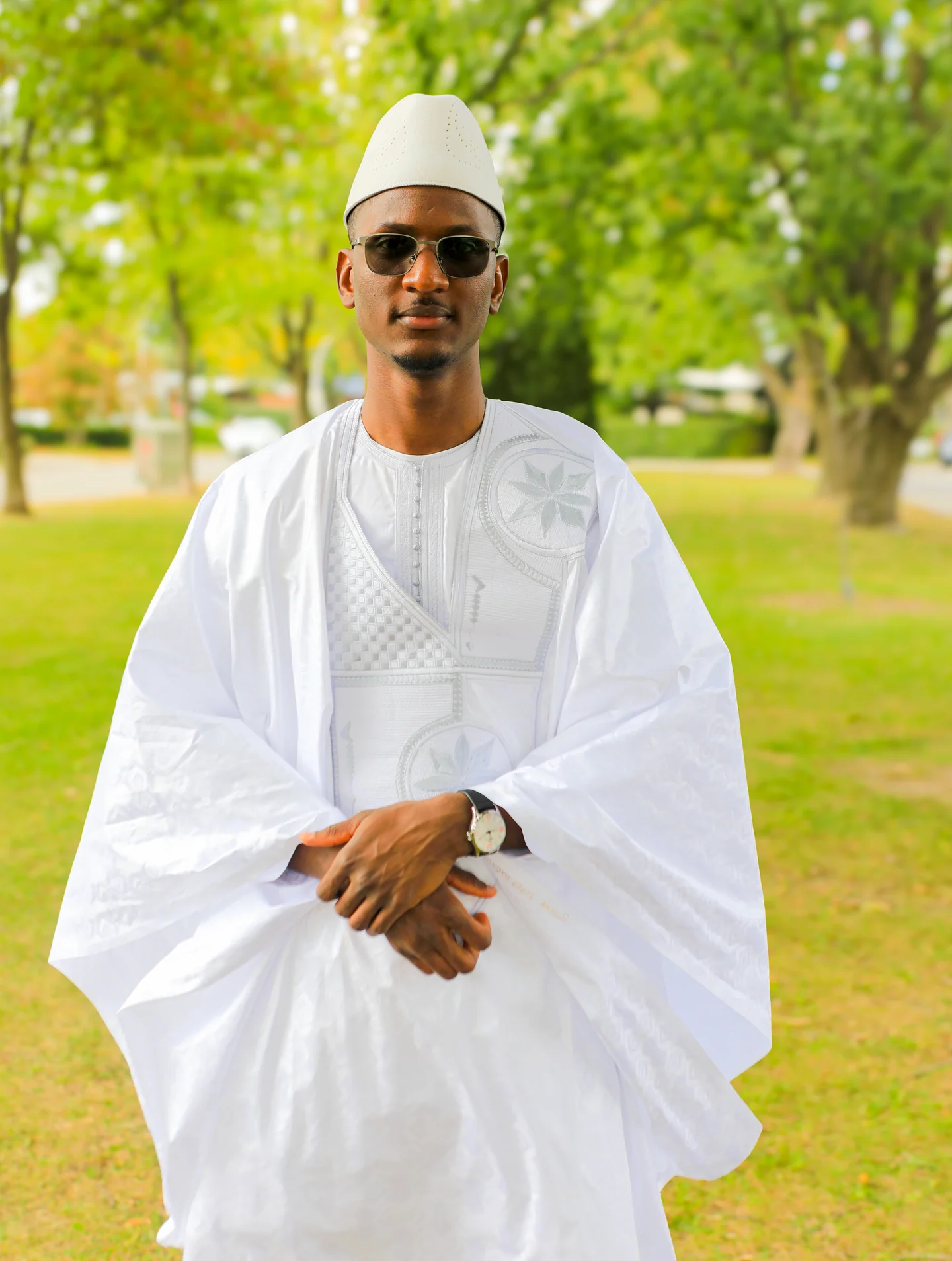 Homme en tenue traditionnelle blanche, chapeau et lunettes de soleil, debout dans un jardin.