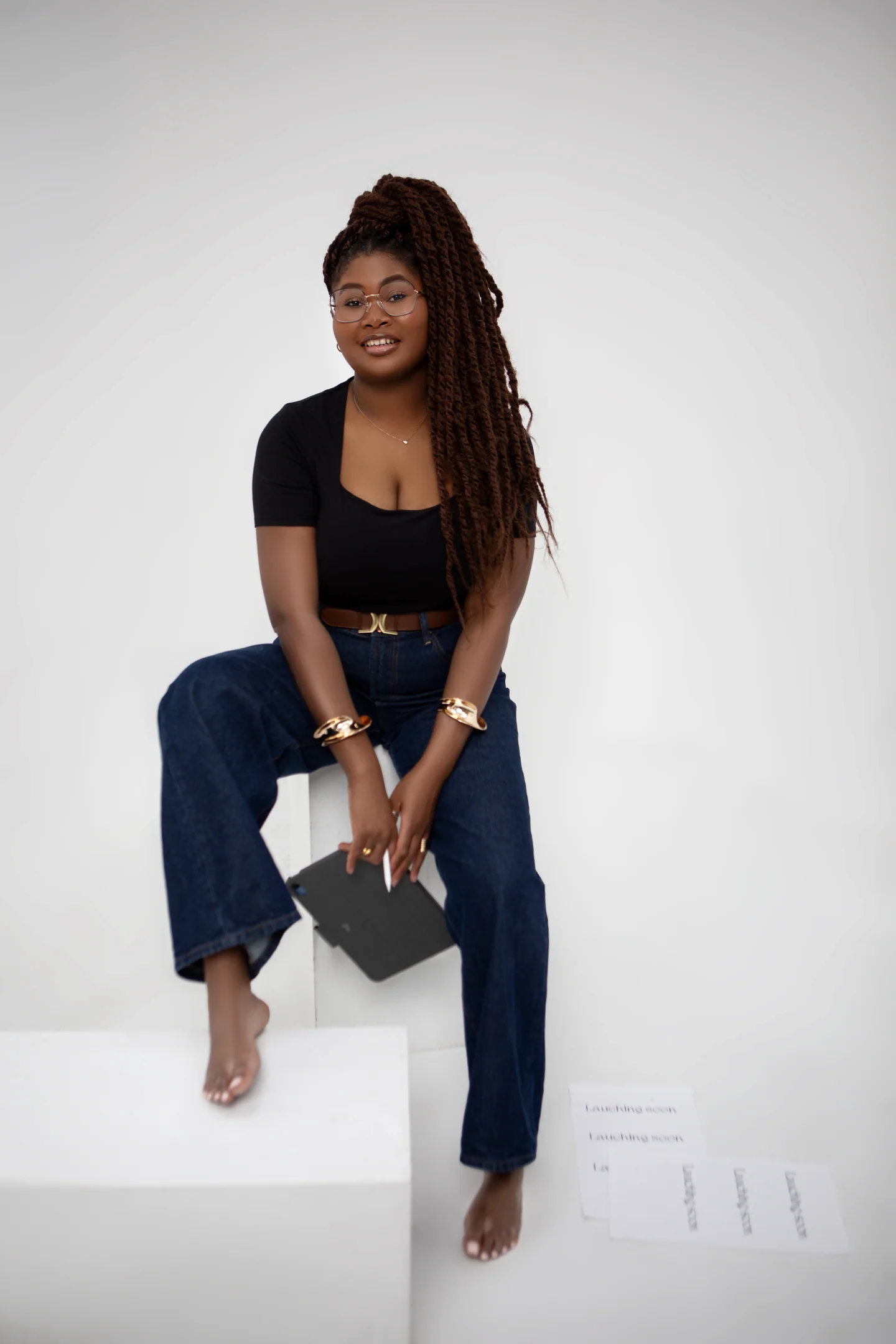 Femme souriante en haut noir et jean, assise sur des cubes blancs avec un carnet dans les mains.