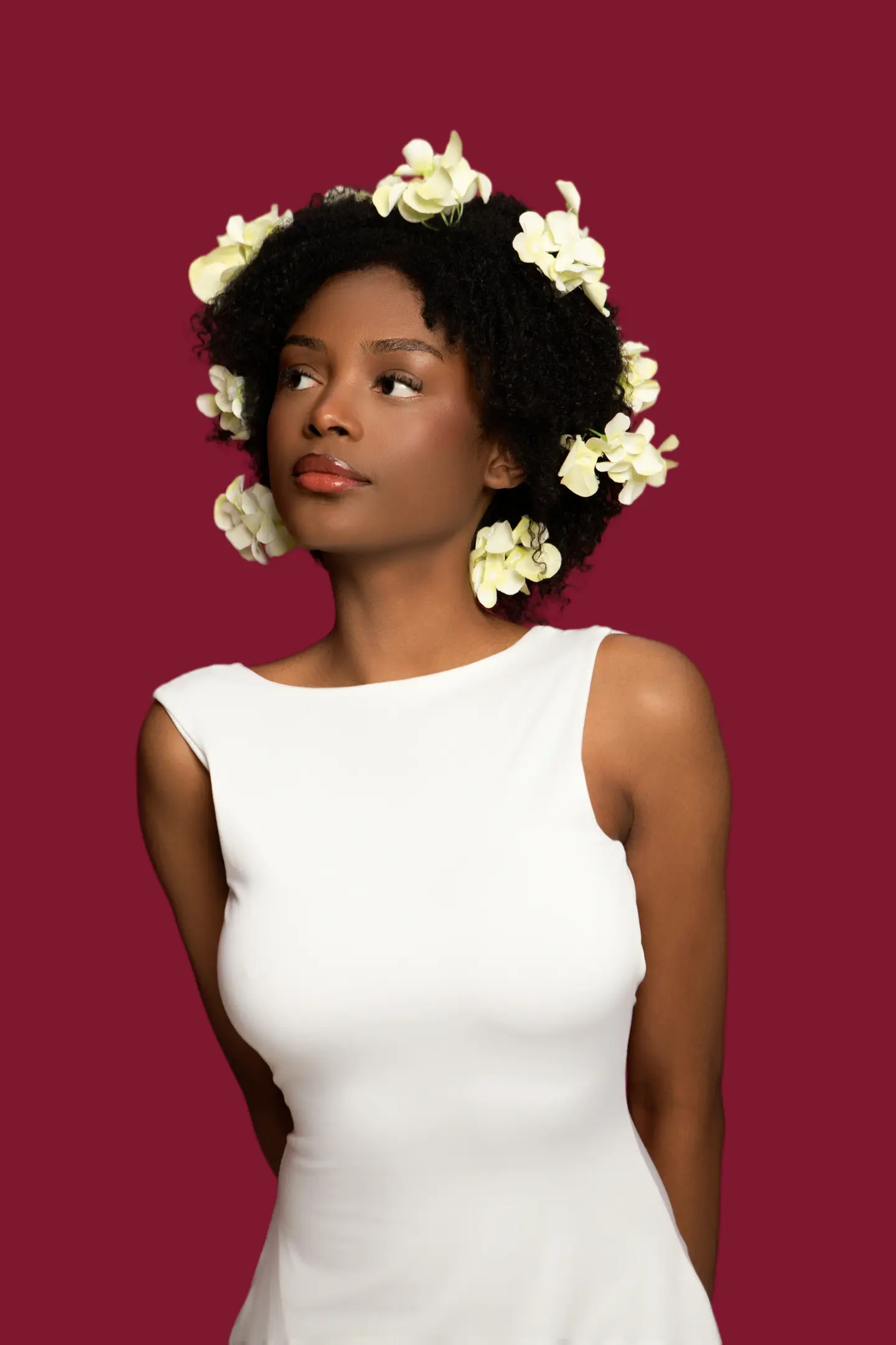 Portrait d’une femme en haut blanc coiffée de fleurs blanches, sur un fond bordeaux.