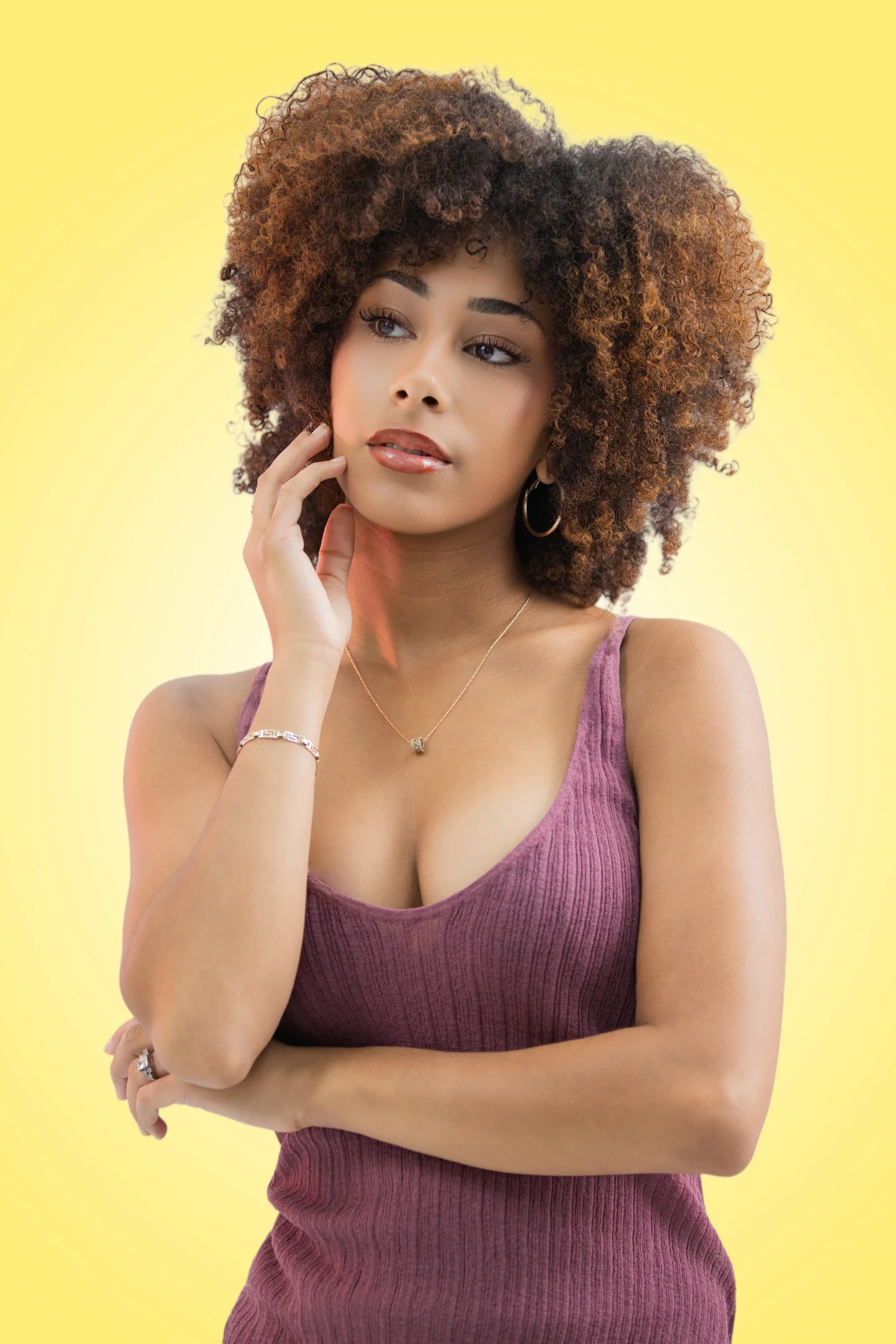 Femme aux cheveux bouclés en robe mauve, une main contre la joue, sur fond jaune.