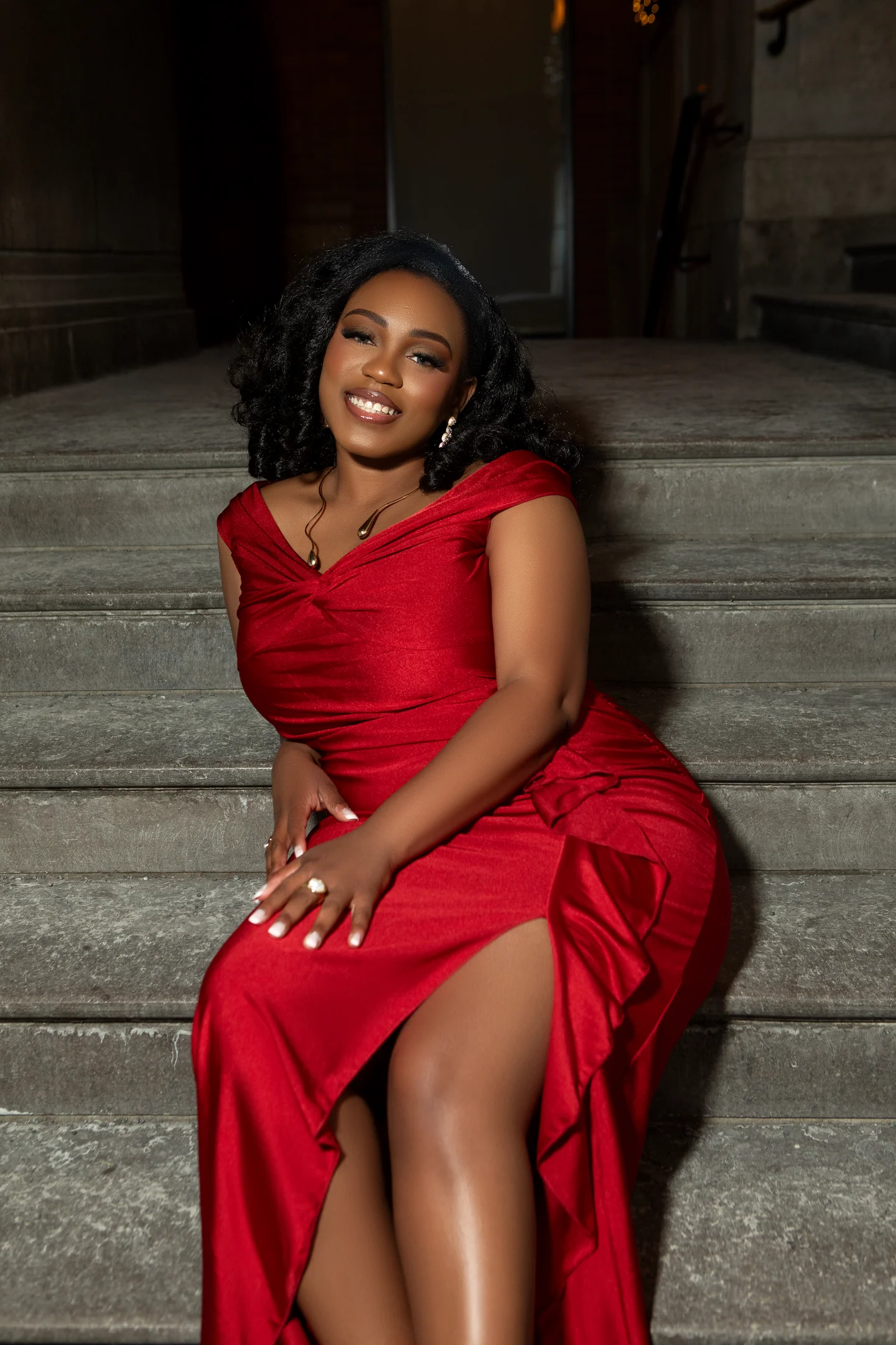 Femme souriante en robe rouge, assise sur un escalier en pierre.