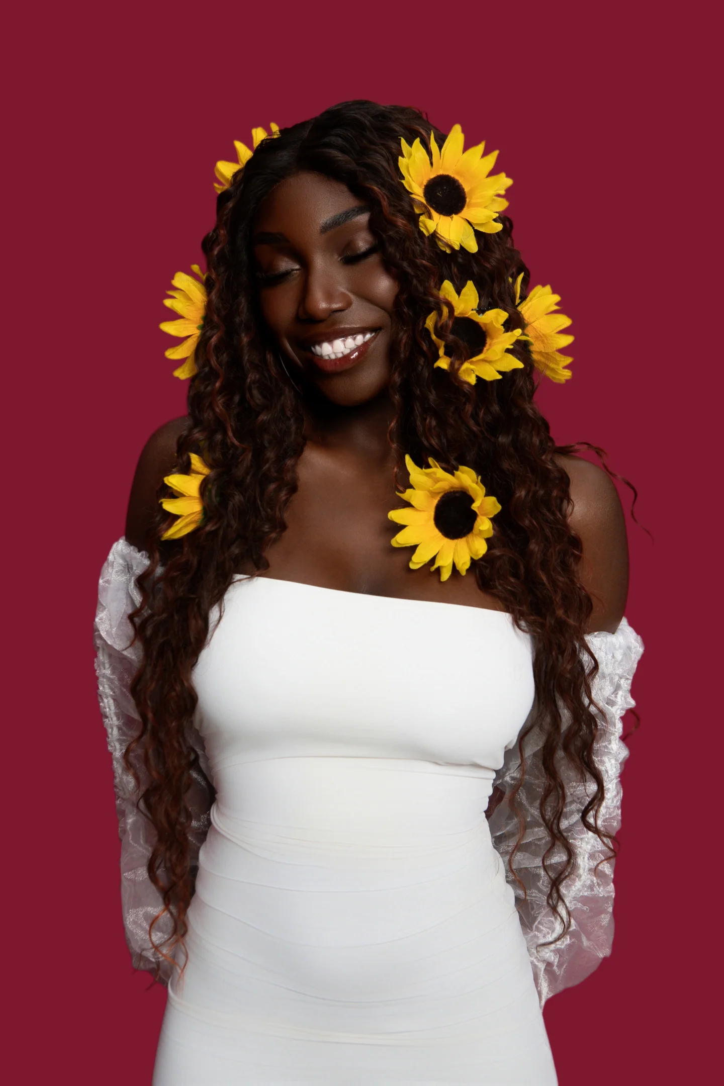 Une femme sourit les yeux baissés, des tournesols dans les cheveux, sur un fond bordeaux.