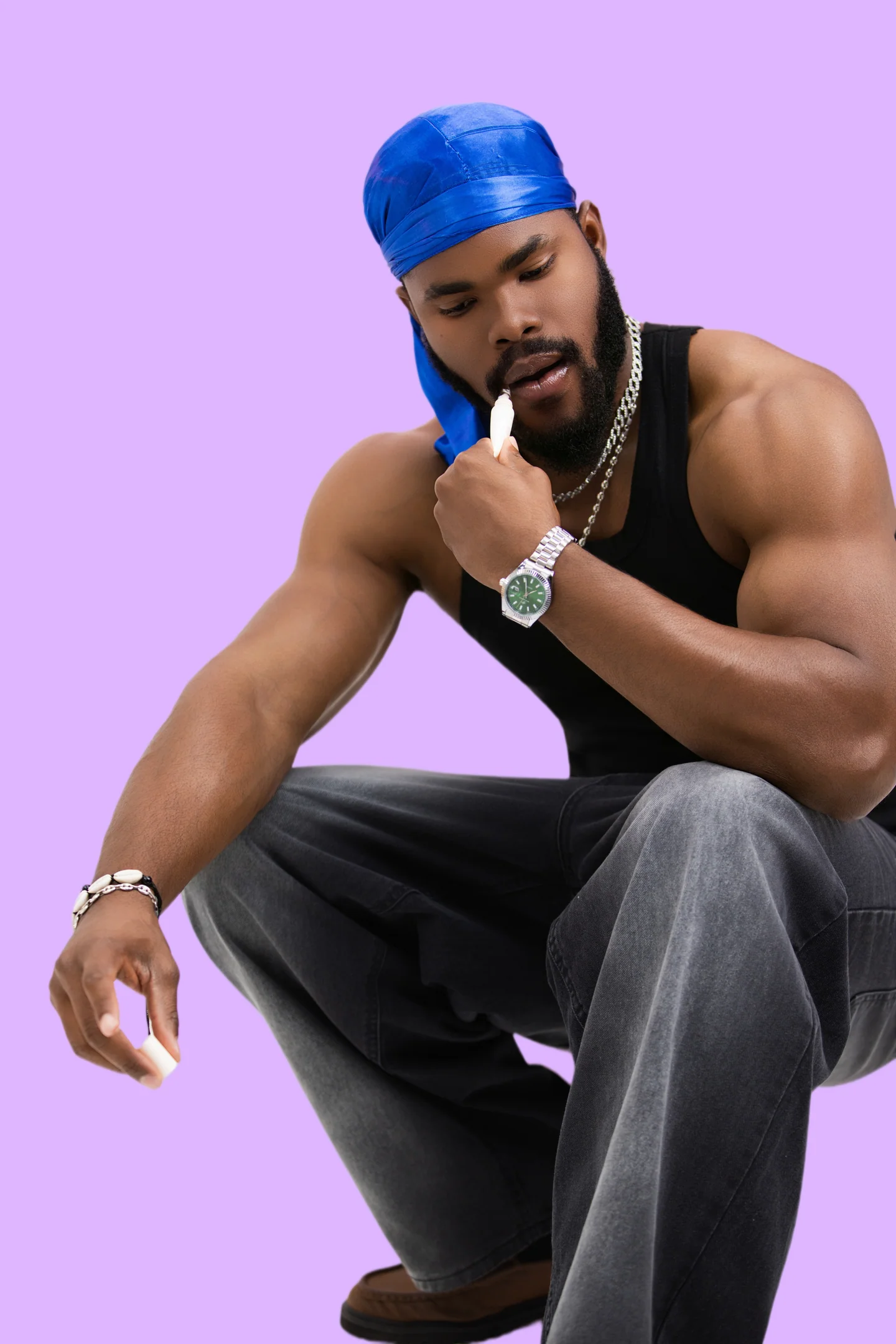 Homme en débardeur noir et durag bleu appliquant un baume à lèvres, assis sur fond violet.
