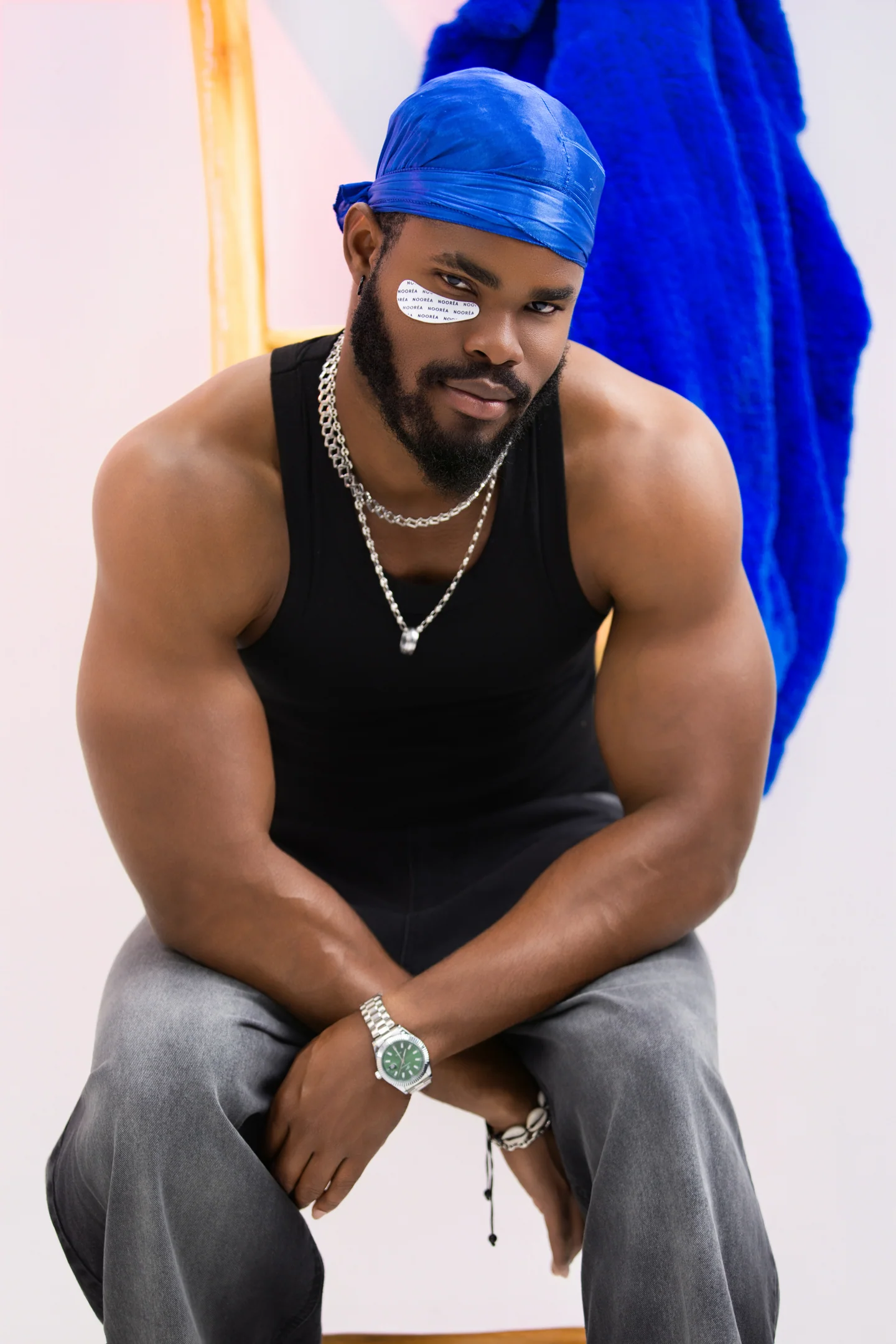 Homme assis en débardeur noir et durag bleu, avec des patchs sous les yeux, devant un peignoir bleu.