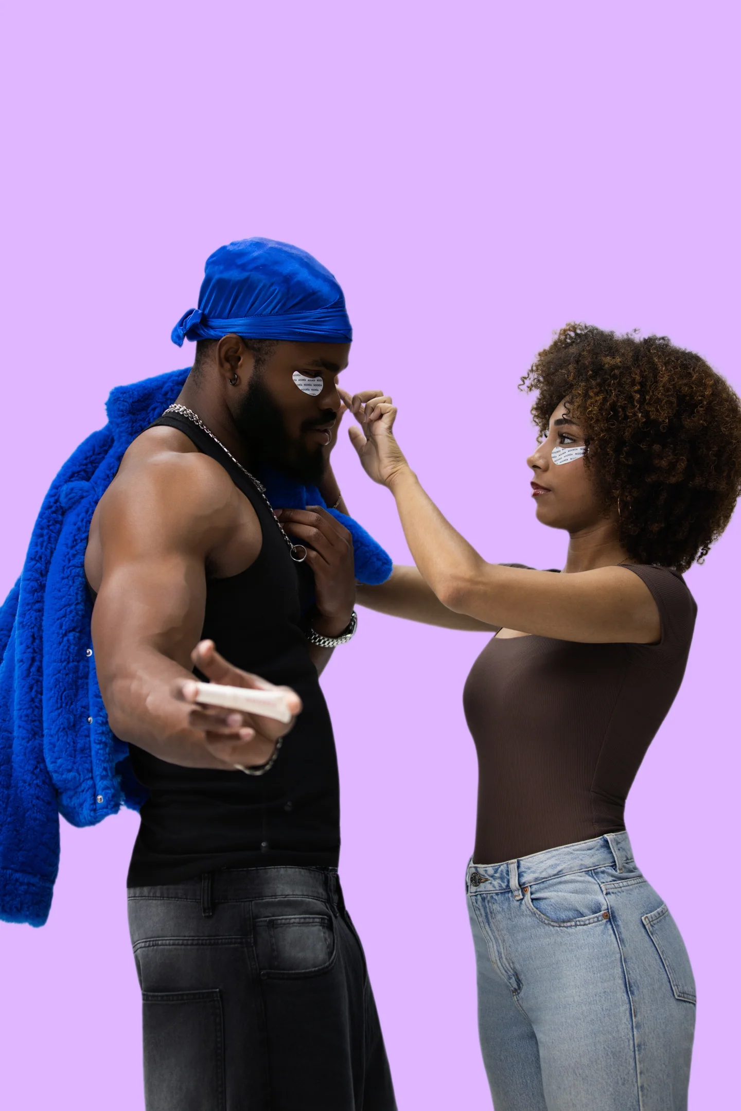 Femme posant un patch sous les yeux d’un homme en durag bleu, tous deux sur fond violet.