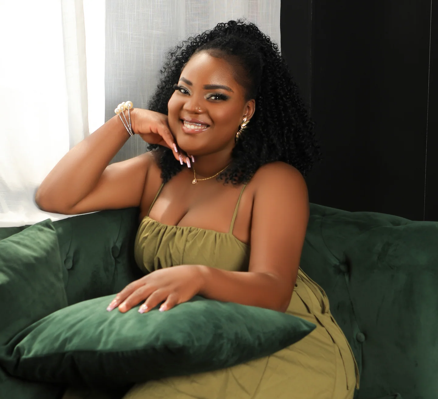 Femme souriante en robe vert olive, assise sur un canapé vert près d’une fenêtre.