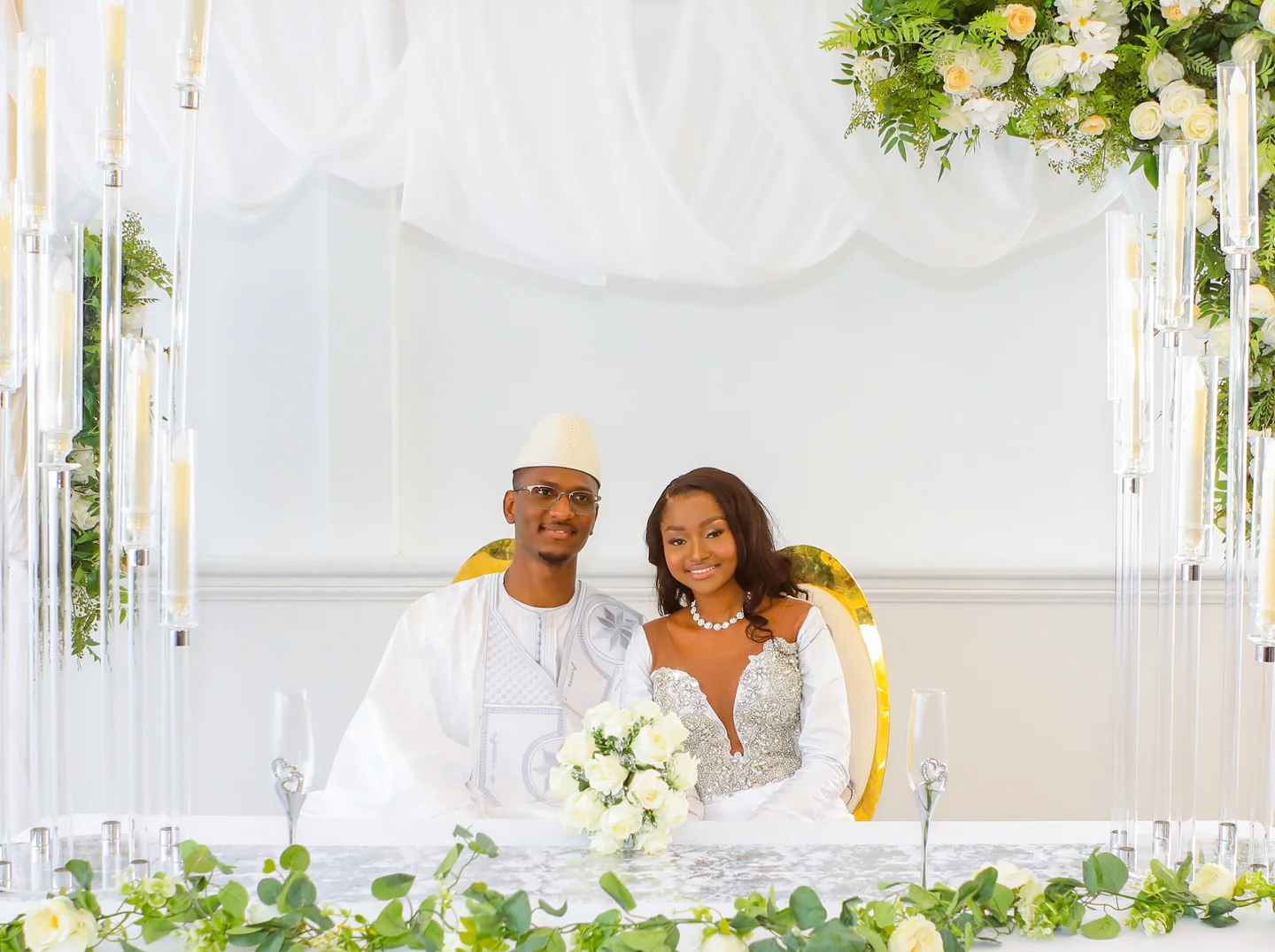 Couple en tenues de mariage blanches, assis derrière une table fleurie sous des voilages blancs.
