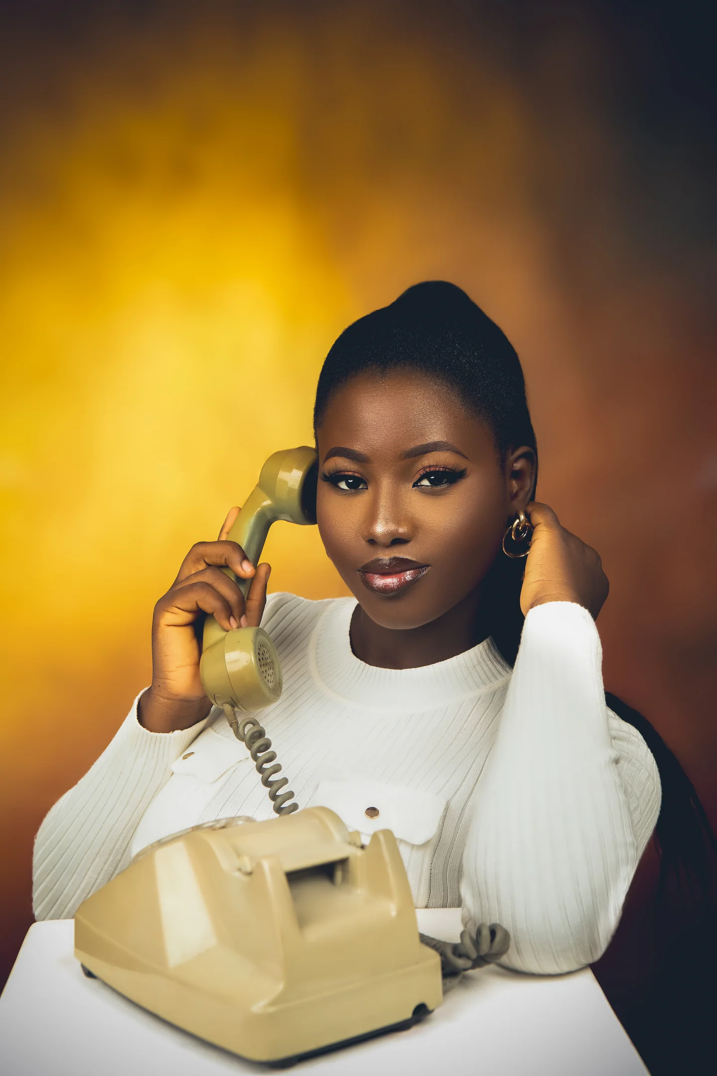 Femme en pull blanc tenant le combiné d’un téléphone crème, éclairée sur un fond jaune doré.